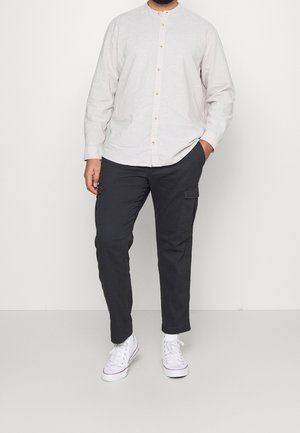 Mężczyzna ubrany w jasnoszarą koszulę na guziki, czarne spodnie cargo i białe wysokie trampki, stojący z jedną ręką w kieszeni.