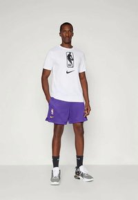 Balta marškinėliai su juodu NBA logo ir Nike ženklu, derinami su purpurinėmis tinklinėmis šortais, ant kurių matyti Nike ir NBA logotipai, ir pilkais sportiniais bateliais.