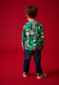 Cămașă verde cu mâneci lungi, cu un print festiv cu animale, asortată cu blugi închisi la culoare și pantofi de o culoare deschisă. Fundal roșu.