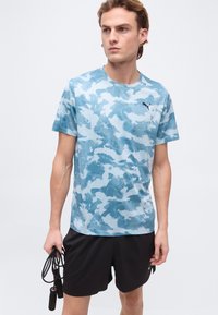 Νεαρός άνδρας που φοράει μπλε καμουφλάζ κοντομάνικο πουκάμισο και μαύρο σορτς, κρατώντας ένα μαύρο σχοινί άλματος στο αριστερό του χέρι, στέκεται μπροστά σε ένα απλό φόντο.