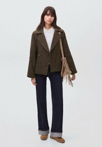 Giacca a doppio petto marrone a quadri con ampi rever, abbinata a un maglione bianco, jeans scuri e scarpe color cuoio; tiene una borsa con frange.