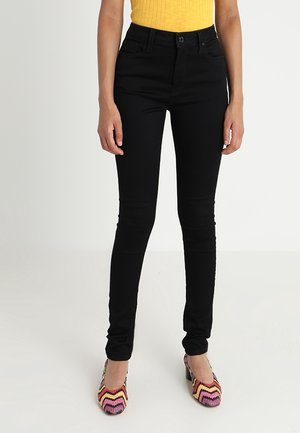 Person trägt schlanke schwarze Jeans, gelbes ärmelloses Oberteil und mehrfarbige, im Zickzackmuster gestaltete High-Heels und steht auf weißem Boden.