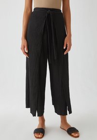 Pantalon large noir texturé avec une ceinture à nouer, présentant un design ouvert à l'avant et une légère drape. Associé à des sandales à glissière tissées.