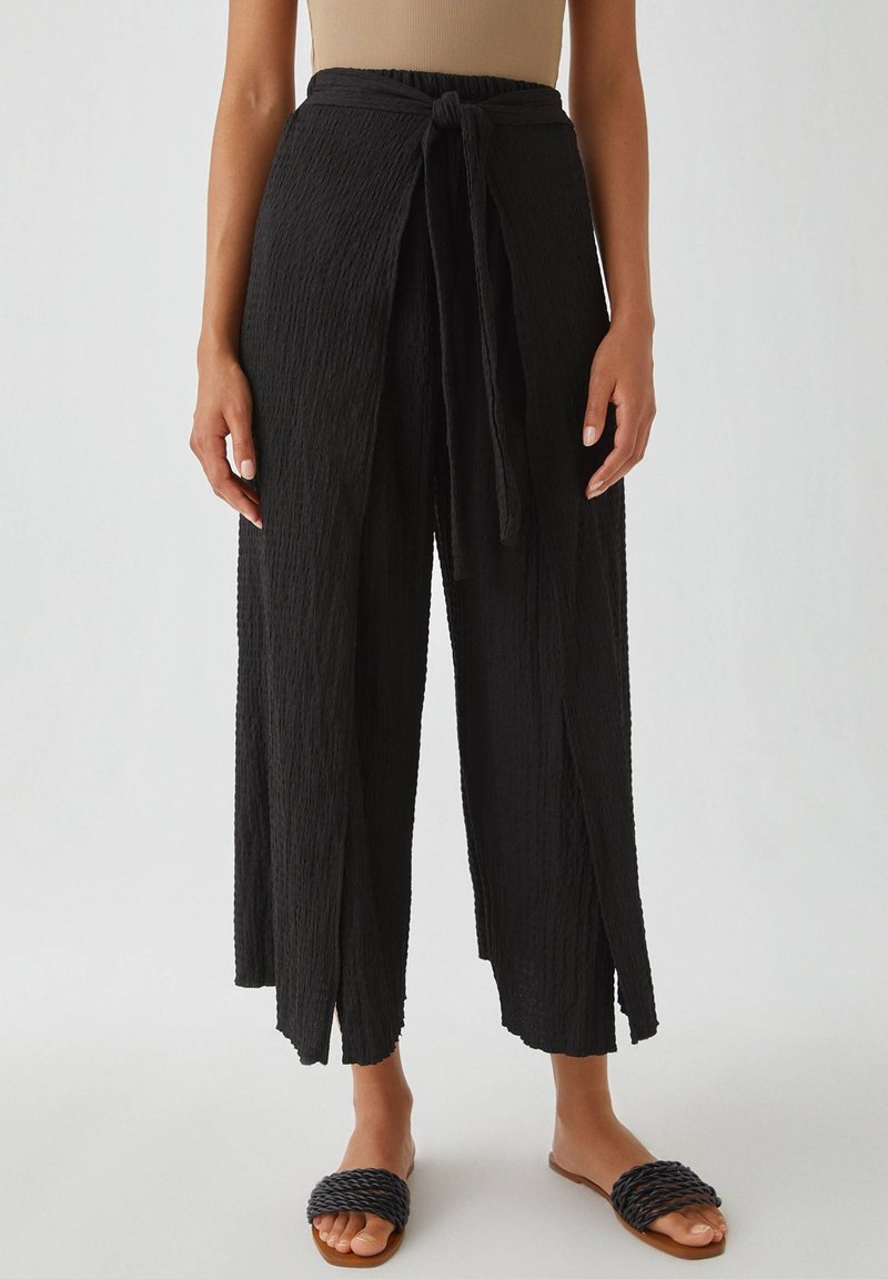 Pantalon large noir texturé avec une ceinture à nouer, présentant un design ouvert à l'avant et une légère drape. Associé à des sandales à glissière tissées.