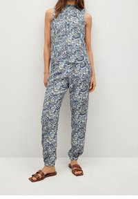 Vrouw in een bijpassende mouwloze top en broek in een blauw en groen paisleyprint, gecombineerd met bruine platte sandalen, staand tegen een effen achtergrond.