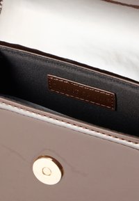 Λαμπερό μπεζ τσάντα χειρός με χρυσό μαγνητικό κούμπωμα, εμφανίζοντας μαύρη υφασμάτινη εσωτερική επένδυση και καφέ δερμάτινη ετικέτα με ανάγλυφο το "Call It Spring."