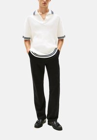 Uomo che indossa una polo bianca a maniche corte con texture e strisce nere, pantaloni neri e mocassini neri con nappine, in piedi con le mani nelle tasche.