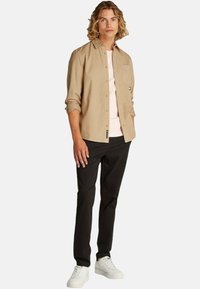Camicia beige a bottoni, t-shirt rosa girocollo sotto, pantaloni neri e sneakers bianche. Presenta una tasca frontale e un'etichetta con logo.
