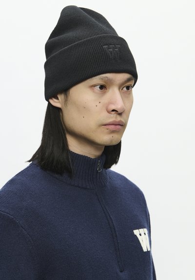 Bonnet noir côtelé avec un revers et un logo en relief. Porté avec un pull en laine bleu marine doté d'un col haut et d'une fermeture zippée quart.