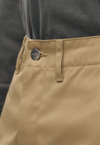 Καφέ παντελόνια με απαλή υφή και κλείσιμο με κουμπί. Διαθέτουν θηλιές για ζώνη και σχεδιασμό με επίπεδη μπροστινή όψη. Το φόντο είναι γκρι.