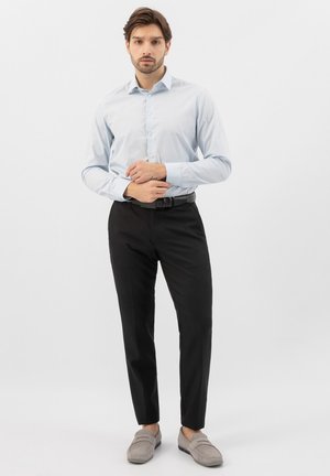 Mann steht vor einem schlichten weißen Hintergrund, trägt ein hellblaues Hemd, schwarze Hose mit schwarzem Gürtel und graue Loafers.