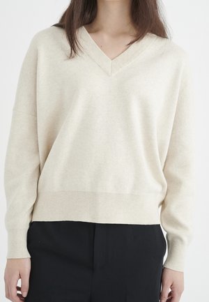 Person iført en cremefarvet V-hals langærmet sweater og sorte bukser mod en ensfarvet hvid baggrund.