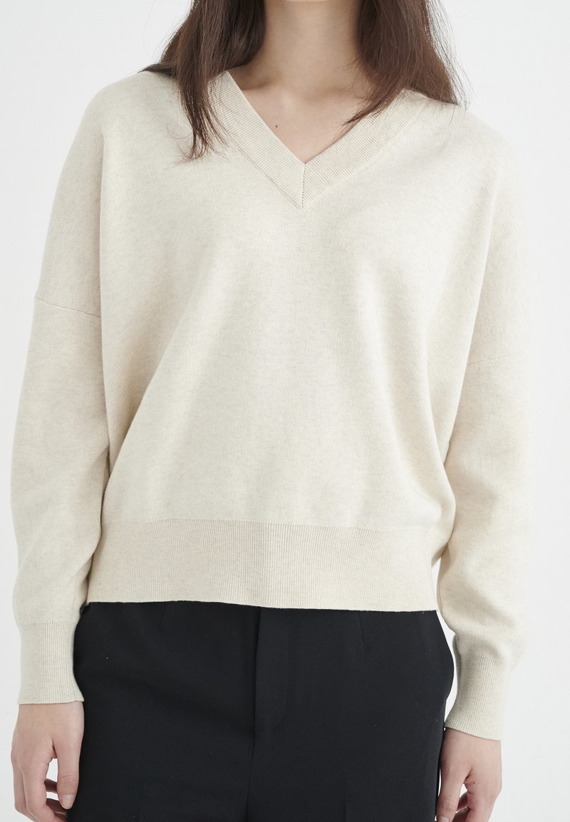 Personne portant un pull crème col en V à manches longues et un pantalon noir sur un fond blanc uni.