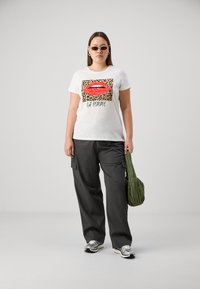 T-shirt blanche avec un graphique de lèvres rouges et un accent de léopard ; associée à un pantalon cargo noir et un sac matelassé vert, complétée par des chaussures de sport.