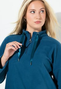 Teal fleece pullover med kvart-lynlås krave, der har teal satin snore og en blød tekstur. Den har en løs pasform og lange ærmer.