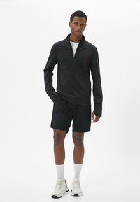 Maglietta nera atletica a maniche lunghe con zip a metà abbinata a pantaloni corti neri. Il modello indossa calze bianche e scarpe da ginnastica chiare, evidenziando un design sporty minimalista.