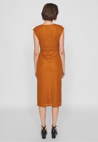 Kleid in verbranntem Orange bis zum Knie mit strukturiertem Rautenmuster, kurzen Ärmeln und taillierter Passform. Getragen mit schwarzen Sandalen mit dicken Absätzen.