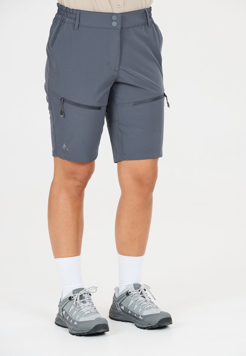 Personne portant un short de randonnée gris mi-long avec des poches zippées sur les cuisses, des chaussettes blanches montantes, et des chaussures de randonnée grises, debout sur fond blanc.