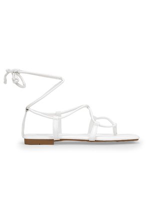 Hvid flad sandal med tynde remme og ankelbind, med tåsløjfe og brun sål.