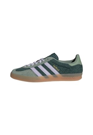 Zelená teniska Adidas Gazelle s bílými pruhy, zlatým nápisem „"GAZELLE"“, bílými tkaničkami, semišovou texturou a gumovou podrážkou, zobrazená z bočního pohledu.