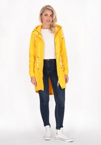 Gul vandtæt jakke med hætte, elastiske manchetter, justerbare snore og en afslappet pasform, parret med mørkeblå jeans og hvide sneakers.