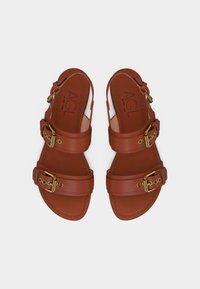 Sandales plates en cuir marron avec deux brides réglables à boucle dorée et bouts ouverts, portant la marque "AGL" à l'intérieur.