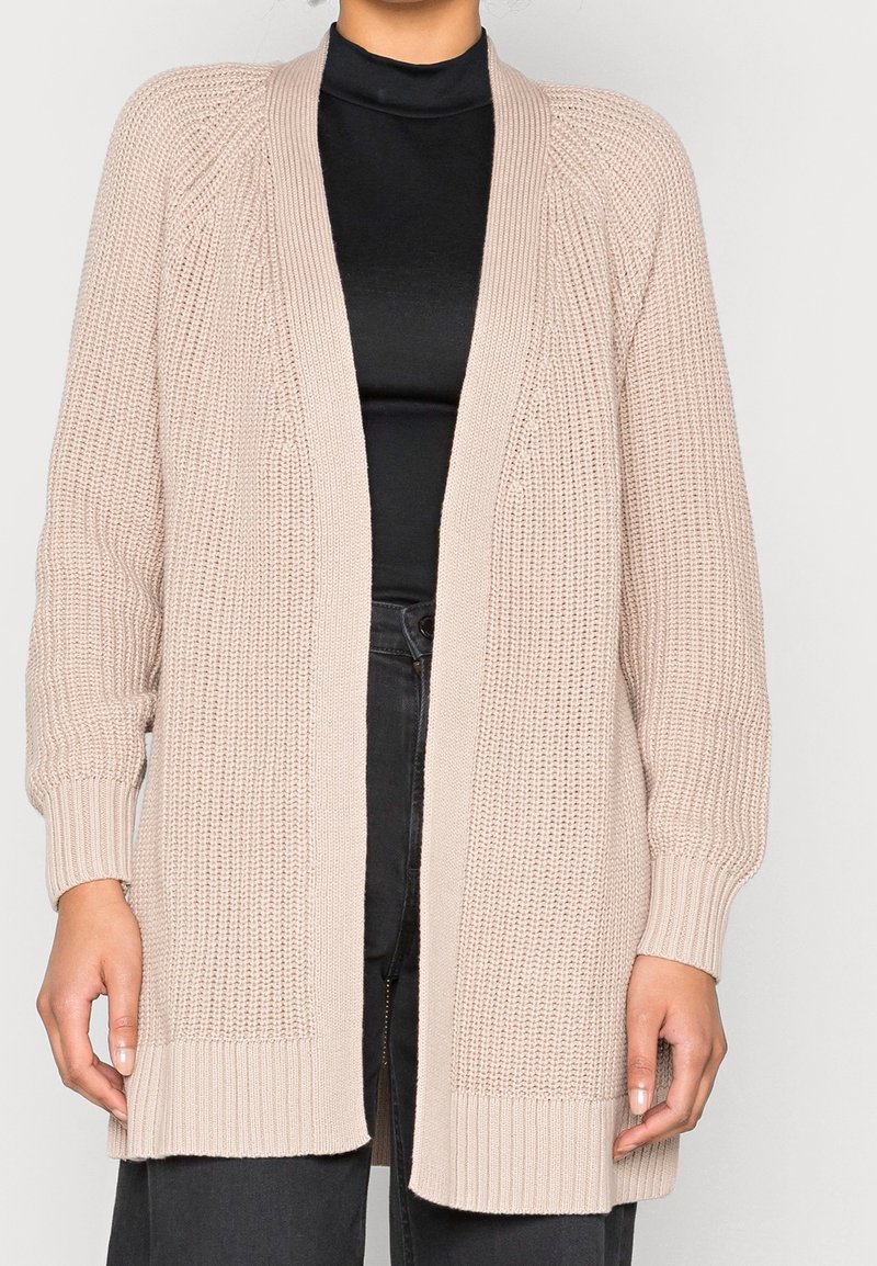 Cardigan en tricot beige à ouverture devant, manches côtelées et variations de texture. Présente une coupe ample et est porté sur un haut noir.