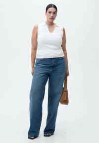 Bela majica bez rukavov z V-izrezom, v kombinaciji z modrimi širokimi denimski hlačami. Model drži rjavo torbico. Enostaven dizajn in lahek material.