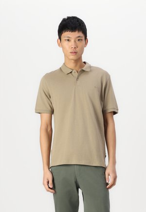Polo shirt kaki en coton, avec un col classique, des manches courtes et un logo discret sur la poitrine. Porté avec un pantalon vert.