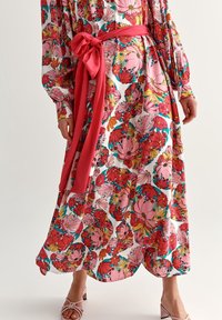Vestido maxi con estampado floral en blanco con vibrantes flores rosas, rojas y amarillas. Incluye un cinturón rosa brillante atado en la cintura y mangas largas.