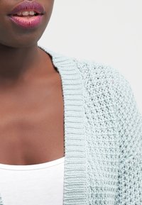 Cárdigan de punto azul claro con textura de tejido abierto, usado sobre una camiseta sin mangas blanca. Presenta un escote acanalado y detalles sutiles.