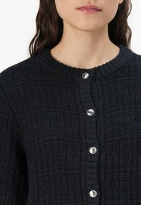 Marineblauwe gebreide cardigan met een gestructureerd patroon, een geribbelde halslijn en vijf zilverkleurige knopen aan de voorkant.