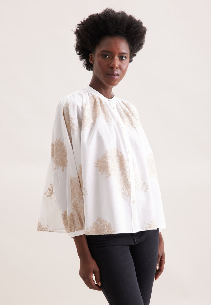 Camicia bianca con bottoni e ricami floreali dorati. Presenta un collo rotondo e maniche voluminose, realizzata in tessuto leggero.