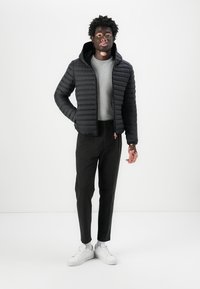 Polstret svart jakke med hette, glidelås foran og ribbestrikkede mansjetter, parret med en grå genser og svarte bukser. Hvite joggesko fullfører looksen.