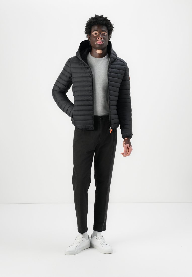 Veste noire matelassée avec capuche, fermée par zip, et poignets côtelés, associée à un pull gris et un pantalon noir. Des baskets blanches complètent le look.