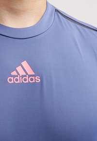 Camiseta deportiva azul hecha de tejido suave. Presenta el logo de Adidas en rosa en la parte frontal y detalles negros en los hombros. Cuello redondo.