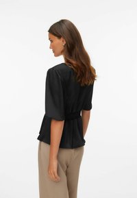 Sort bluse med korte, puffede ermer, innsnevret midje og jevn tekstur. Brukt med beige bukser, som viser en slank silhuett.