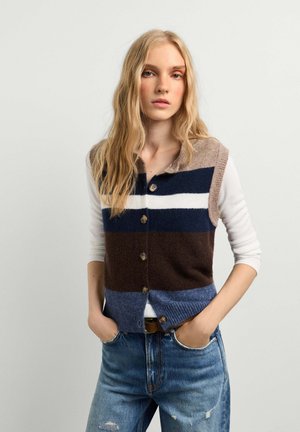 Young woman with long blonde hair wearing a buttoned striped knit vest over a white top and blue jeans with hands in pockets.