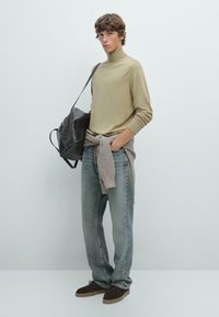 Ljus olivgrön polotröja, avslappnade grå jeans och bruna mockaskor; håller en svart axelväska med en knuten beige tröja i midjan.