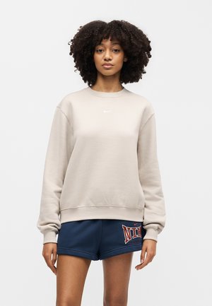 Młoda kobieta o kręconych włosach ubrana w jasnobeżową bluzę Nike i granatowe spodenki Nike, stojąca na jednolitym tle.