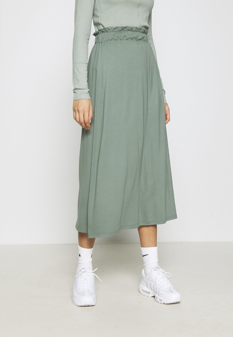 Femme portant une jupe midi vert sauge avec une taille élastique, un haut côtelé à manches longues vert clair, des chaussettes blanches Nike et des baskets blanches.