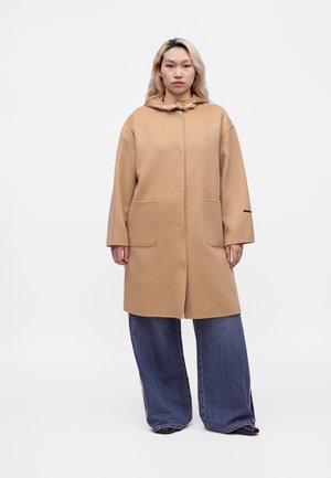 Personne aux longs cheveux clairs portant un manteau à capuche beige et un jean bleu large, debout devant un fond uni.