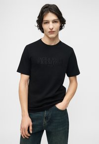 Νεαρός άνδρας με μεσαίου μήκους σκούρα μαλλιά, φορώντας μαύρο T-shirt "Jack & Jones Premium" και μπλε τζιν, με το ένα χέρι στην τσέπη.