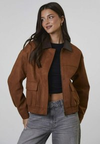 Młoda kobieta z długimi ciemnymi włosami, ubrana w brązową kurtkę na czarny top crop i szare dżinsy, patrzy na bok z lekkim uśmiechem.