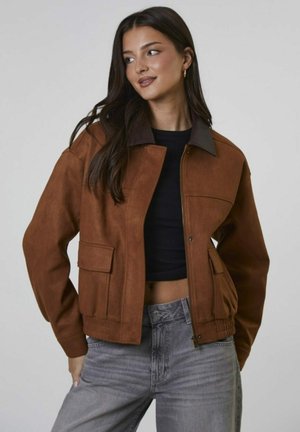 Jonge vrouw met lang, donker haar, gekleed in een bruine jas over een zwarte crop top en grijze jeans, kijkt zijwaarts met een lichte glimlach.