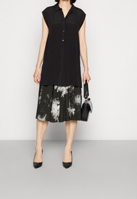 Kvinna i ärmlös svart skjorta med knappar, svart och vit tie-dye kjol, svarta spetsiga klackar, håller en svart handväska vid sin sida.