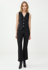 Czarna bezrękawnik z dekoltem w serek i pięcioma złotymi guzikami, w zestawie z czarnymi spódnicami dzwonowymi i czarnymi butami na obcasie ze spiczastym noskiem. Gładka tekstura.