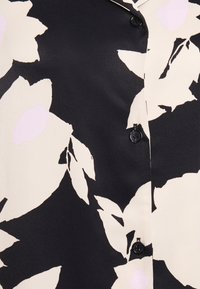 Hemd mit Knöpfen auf schwarzem Hintergrund, das große cremefarbene Blumen mit pinken Akzenten zeigt. Glatte Textur; ein schwarzer Knopf sichtbar.