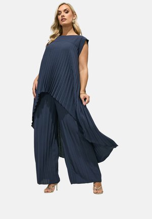 Vrouw draagt een marineblauwe plissé mouwloze tuniek met asymmetrische zoom en bijpassende broek met wijde pijpen, gecombineerd met gouden sandalen met bandjes.