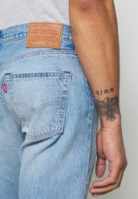 Jeans Levi's en un azul claro deslavado con un parche de cuero en la cinturilla, que muestran detalles de costura, una etiqueta roja en el bolsillo trasero y un tatuaje visible.